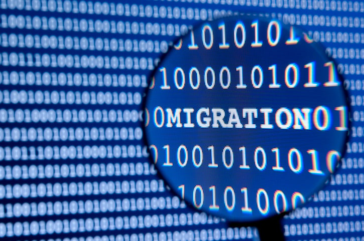 A magnifying glass in front of an illuminated blue tinted computer screen full of rows of programming code with the word migration.